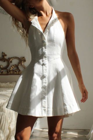 Woman in a white dress standing in a sunlit bedroom with checkered floor and ornate bed.